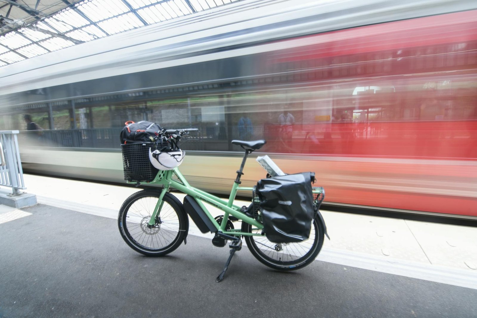 A commuter e-bike locked to a city bike rack with a pannier bag and grocery tote